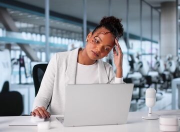 A stressed-out woman, holding her head while facing a laptop, in a modern office environment.