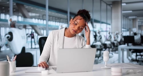 A stressed-out woman, holding her head while facing a laptop, in a modern office environment.