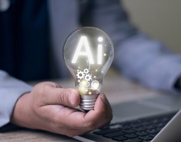 A businessperson holding a glowing light bulb, with the letters "Ai" and gears inside the light bulb, representing technology, artificial intelligence, and machinery.