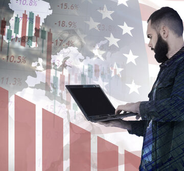Concept image of economic fluctuations in the U.S., featuring a man analyzing financial trends on a laptop against a background of the American flag and declining stock graphs.