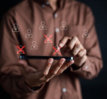 Conceptual image of corporate restructuring, mass layoffs, or employee downsizing showing a person using a mobile device to manage a workforce database using red "X" icons symbolize the selection of specific personnel for termination or restructuring.