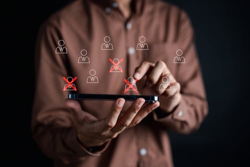 Conceptual image of corporate restructuring, mass layoffs, or employee downsizing showing a person using a mobile device to manage a workforce database using red "X" icons symbolize the selection of specific personnel for termination or restructuring.