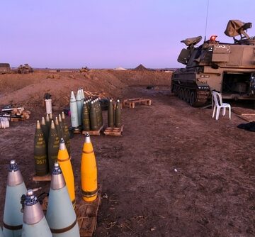 An Israeli Defense Forces (IDF) M109 howitzer artillery system stationed near the Gaza Strip border with 155mm projectiles stacked in various colors indicating different types of shells.
