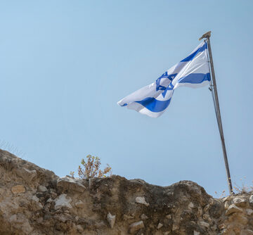 The Israel flag flying on a flagpole atop a stone wall.