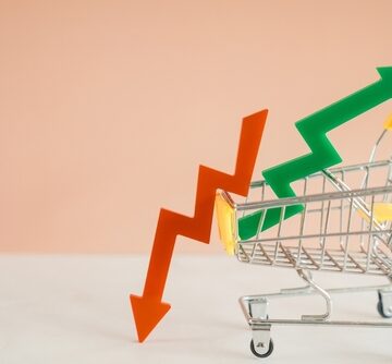 Concept illustration of a shopping cart carrying a falling red arrow and a rising green arrow, likely representing economic concepts such as inflation, market volatility, or changing consumer prices.