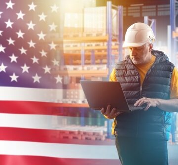 A warehouse worker in a hard hat holding a laptop in front of a superimposed American flag, representing a U.S.-based industrial or logistics company.