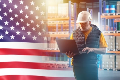 A warehouse worker in a hard hat holding a laptop in front of a superimposed American flag, representing a U.S.-based industrial or logistics company.