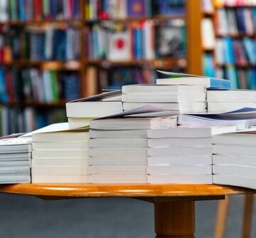A,Large,Pile,Of,Paperback,And,Hardcover,Books,Sits,On A large collection of books stacked on a wooden table in a library or bookstore.