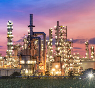 An oil refinery and petrochemical plant illuminated at twilight, featuring characteristic industrial components like distillation towers, complex piping networks, and storage infrastructure.