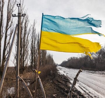 A tattered Ukrainian flag is displayed on a makeshift pole beside a muddy road, in the Donetsk region of Ukraine.