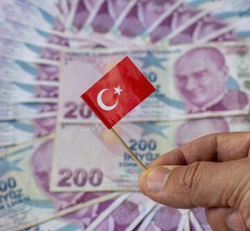 A circle of Turkish Lira banknotes, with a hand holding a miniature Turkish flag above them.