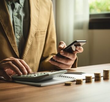 A businessman using a phone and calculator in front of stacks of coins.