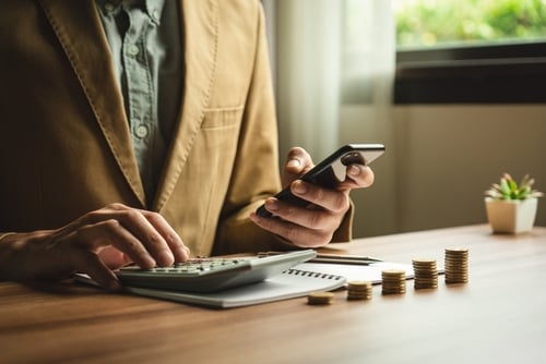 Businessman,Calculates,Investment,Income,,Financial,Business,Investments,Analyze,Risks,,Research A businessman using a phone and calculator in front of stacks of coins.