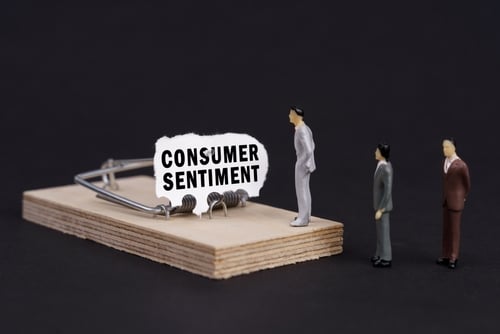 Miniature businessmen cautiously approaching a mousetrap baited with "Consumer Sentiment," illustrating the current economic anxiety as major sentiment indicators reach historic lows.