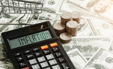 Economic inflation concept showing a calculator displaying the word "INFLATION" resting on a pile of US dollar bills, accompanied by stacked coins and a miniature shopping cart.