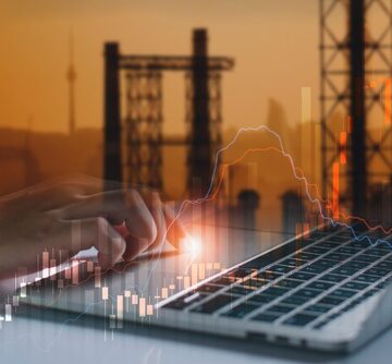 A laptop with stock market candlestick graphs superimposed over silhouettes of oil pumpjacks and refinery towers.