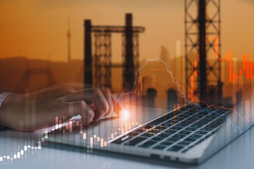 Professional,Analyzing,Global,Energy,Crisis,And,Oil,Price,Spikes.,Oil A laptop with stock market candlestick graphs superimposed over silhouettes of oil pumpjacks and refinery towers.