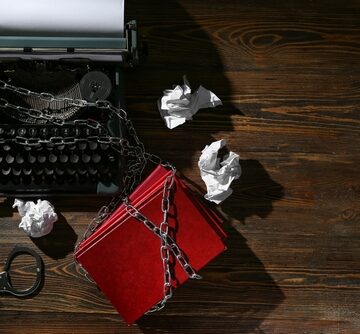 A vintage typewriter and books are bound in heavy metal chains, symbolizing restricted knowledge and the suppression of free speech.