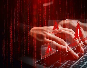 A person typing on a laptop with red, holographic "Cyber Attack" alerts and exclamation marks overlaid on the screen.