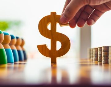 A hand placing a wooden dollar sign between stacked coins and a line of stylized figures. A hand placing a wooden dollar sign between stacked coins and a line of stylized figures.