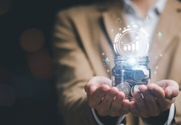 A man holding a jar of coins with a digital overlay of a growth graph icon rising from the jar. A man holding a jar of coins with a digital overlay of a growth graph icon rising from the jar.