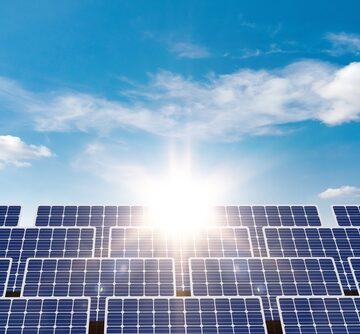 A large array of photovoltaic solar panels positioned under a bright, sunny blue sky with scattered white clouds.