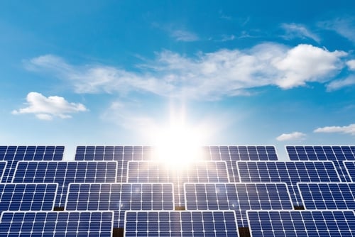 A large array of photovoltaic solar panels positioned under a bright, sunny blue sky with scattered white clouds. A large array of photovoltaic solar panels positioned under a bright, sunny blue sky with scattered white clouds.