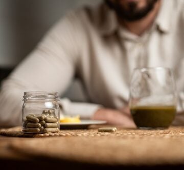A person sits in front of a glass jar filled with green capsules alongside a glass containing a green liquid which appears to be the herbal product kratom.
