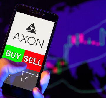 A person's hand holding a mobile phone with the Axon Enterprise logo above green "BUY" and red "SELL" buttons. A person's hand holding a mobile phone with the Axon Enterprise logo above green "BUY" and red "SELL" buttons.