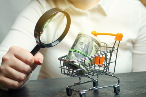 A magnifying glass examining a roll of U.S. currency inside a miniature shopping cart. A magnifying glass examining a roll of U.S. currency inside a miniature shopping cart.