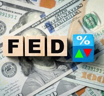Wooden blocks spelling "FED" next to a block with a percentage sign and up-and-down arrows, placed on U.S. $100 dollar bills.