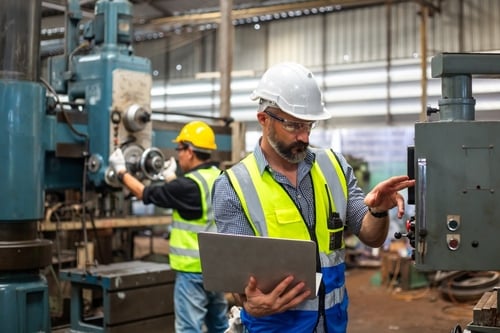 This image shows industrial workers wearing bright yellow safety vests and hard hats while operating machinery and reviewing data on a laptop. This image shows industrial workers wearing bright yellow safety vests and hard hats while operating machinery and reviewing data on a laptop.