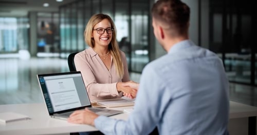 Two professionals shaking hands across a table during a meeting.