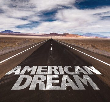 The phrase "AMERICAN DREAM" spray-painted in white stencil lettering on the asphalt of a straight road stretching into a desert landscape.