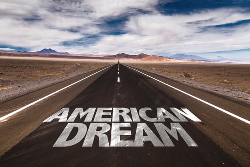 The phrase "AMERICAN DREAM" spray-painted in white stencil lettering on the asphalt of a straight road stretching into a desert landscape. The phrase "AMERICAN DREAM" spray-painted in white stencil lettering on the asphalt of a straight road stretching into a desert landscape.