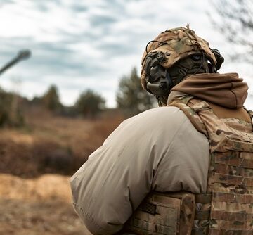 A soldier wearing a camouflage helmet and bulletproof vest with the blurred barrel of a self-propelled artillery mount in the background.