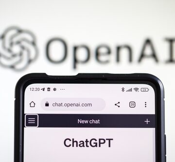 A smartphone displaying the official mobile web browser interface for ChatGPT, set against a background showing the blurred OpenAI logo. A smartphone displaying the official mobile web browser interface for ChatGPT, set against a background showing the blurred OpenAI logo.