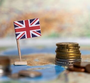 British economy financial system and international trade concept featuring a small British flag toothpick placed next to stacks of coins on a blurred map background. British economy financial system and international trade concept featuring a small British flag toothpick placed next to stacks of coins on a blurred map background.