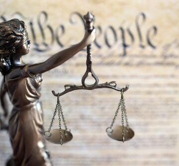The bronze statue of Lady Justice holding scales, positioned in front of a document resembling the U.S. Constitution. The bronze statue of Lady Justice holding scales, positioned in front of a document resembling the U.S. Constitution.