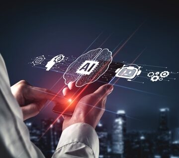 A digital brain icon labeled "AI" holographically displaying interconnected elements like robots and gears, displayed from a mobile device being held by a person. A digital brain icon labeled "AI" holographically displaying interconnected elements like robots and gears, displayed from a mobile device being held by a person.