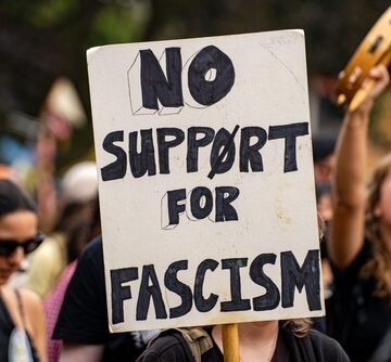 A person in a crowd of protesters holding a sign that reads "NO SUPPORT FOR FASCISM." A person in a crowd of protesters holding a sign that reads "NO SUPPORT FOR FASCISM."