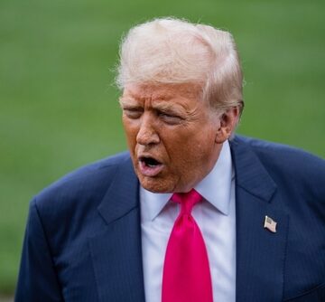 Donald Trump speaking outdoors, wearing a navy blue suit jacket, a white collared shirt, a bright pinkish-red tie, and an American flag lapel pin. Donald Trump speaking outdoors, wearing a navy blue suit jacket, a white collared shirt, a bright pinkish-red tie, and an American flag lapel pin.