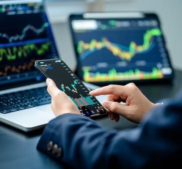 An investor monitoring financial candlestick charts across multiple devices, including a smartphone, a laptop, and a tablet. TRUMP SEES WEALTH BALLOON IN SECOND TERM, DRIVEN BY CRYPTO