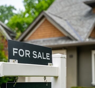 Close-up of a "For Sale" sign positioned on a white post in front of a residential house. Close-up of a "For Sale" sign positioned on a white post in front of a residential house.