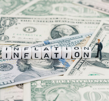 A miniature figure stands next to white cubes spelling "INFLATION" which are stacked on top of U.S. dollar bills. A miniature figure stands next to white cubes spelling "INFLATION" which are stacked on top of U.S. dollar bills.
