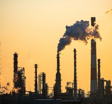 The silhouette of an oil refinery with multiple smokestacks emitting smoke into the atmosphere.