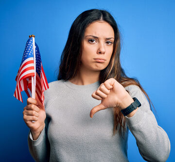 A young woman, with an unhappy face and showing a thumbs down, holding a USA flag with a blue background. A young woman, with an unhappy face and showing a thumbs down, holding a USA flag with a blue background.
