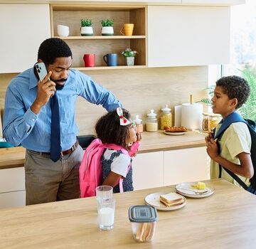 A father wearing a blue button-down shirt and tie while talking on his smartphone in the kitchen and tending to his kids. A father wearing a blue button-down shirt and tie while talking on his smartphone in the kitchen and tending to his kids.