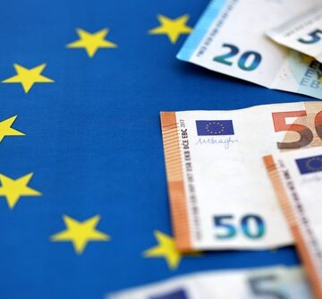 Various euro banknotes (such as 5, 20, and 50 euro notes) placed on top of the blue and yellow flag of the European Union. Various euro banknotes (such as 5, 20, and 50 euro notes) placed on top of the blue and yellow flag of the European Union.
