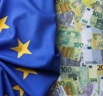 Various denominations of Euro banknotes (€20, €100, €200, €500) spread out alongside the European Union flag. Various denominations of Euro banknotes (€20, €100, €200, €500) spread out alongside the European Union flag.